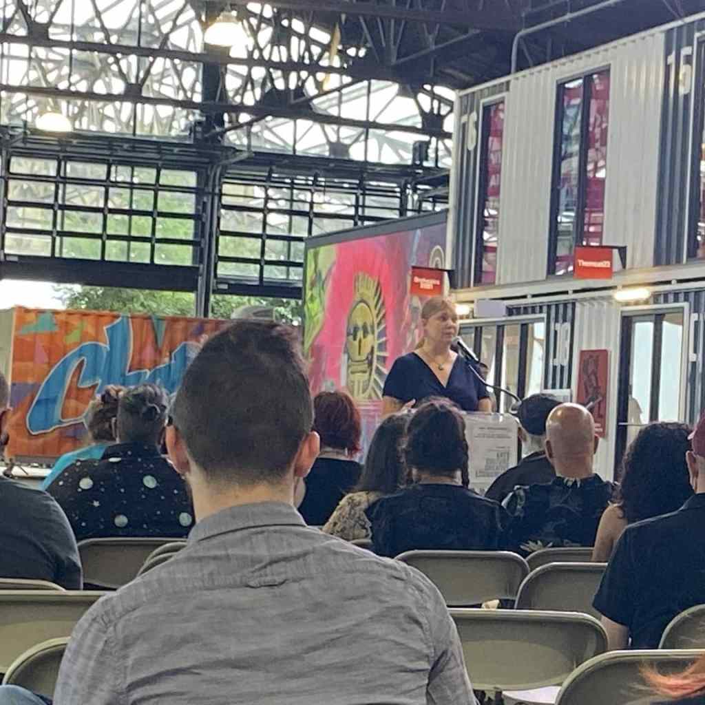 The blog author is wearing a blue dress and reading at a podium in front of a crowd in a high-ceilinged industrial space. 